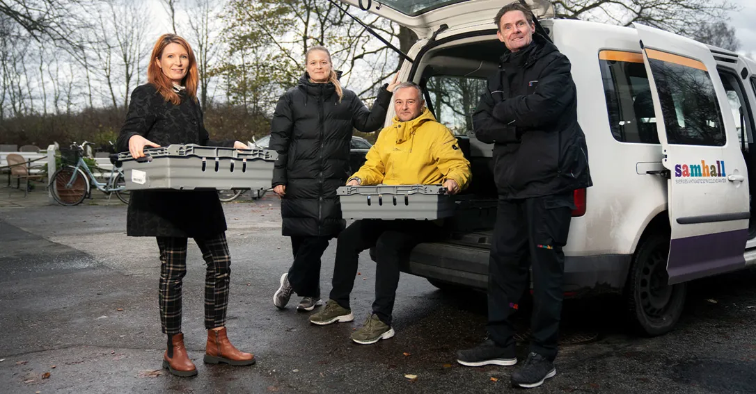 Fyra personer står vid en vit servicebil med bakluckan öppen. Två av dem håller grå plastlådor, en person sitter på bilens lastkant i gul jacka och en annan står med armarna i kors. På bilen syns logotypen ”Samhall”. I bakgrunden finns träd, cyklar och en asfalterad yta utomhus.