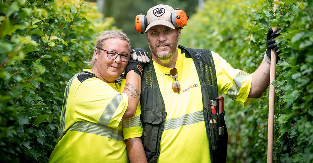 Två personer i arbetskläder står vid en häck och kollar in i kameran