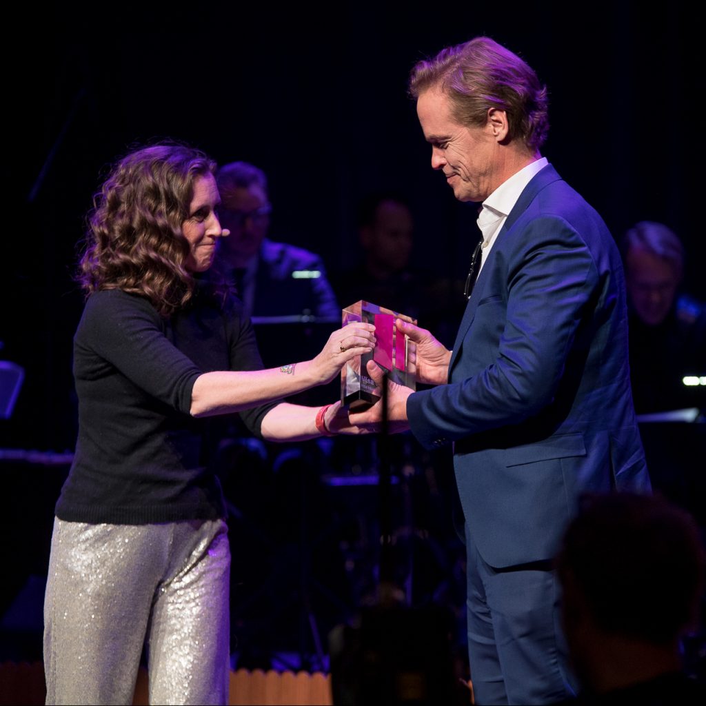 En kvinna och en man står på en scen med mörk bakgrund och scenbelysning. De håller tillsammans i en glas- eller akrylpokal som överlämnas mellan dem, medan musiker syns suddigt i bakgrunden.