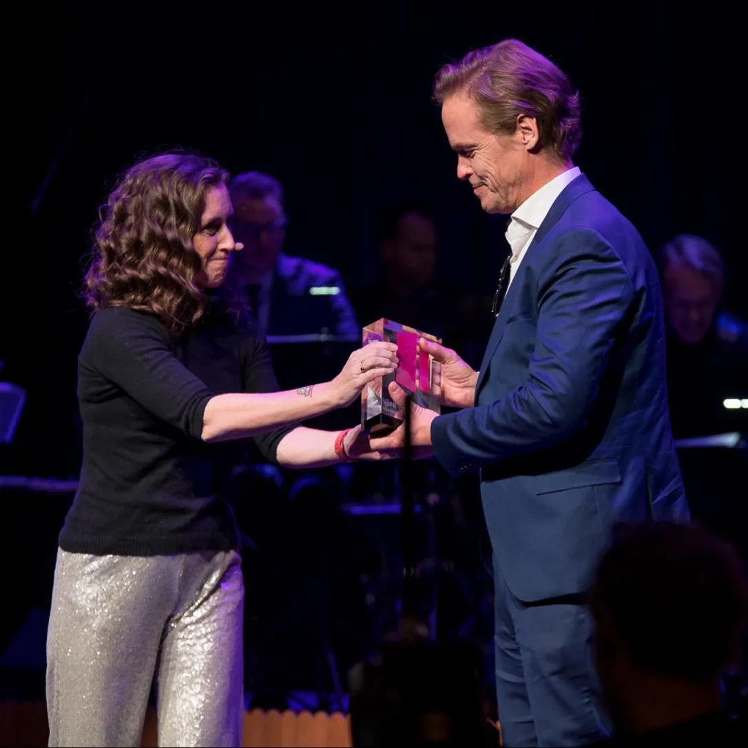 En kvinna och en man står på en scen med mörk bakgrund och scenbelysning. De håller tillsammans i en glas- eller akrylpokal som överlämnas mellan dem, medan musiker syns suddigt i bakgrunden.