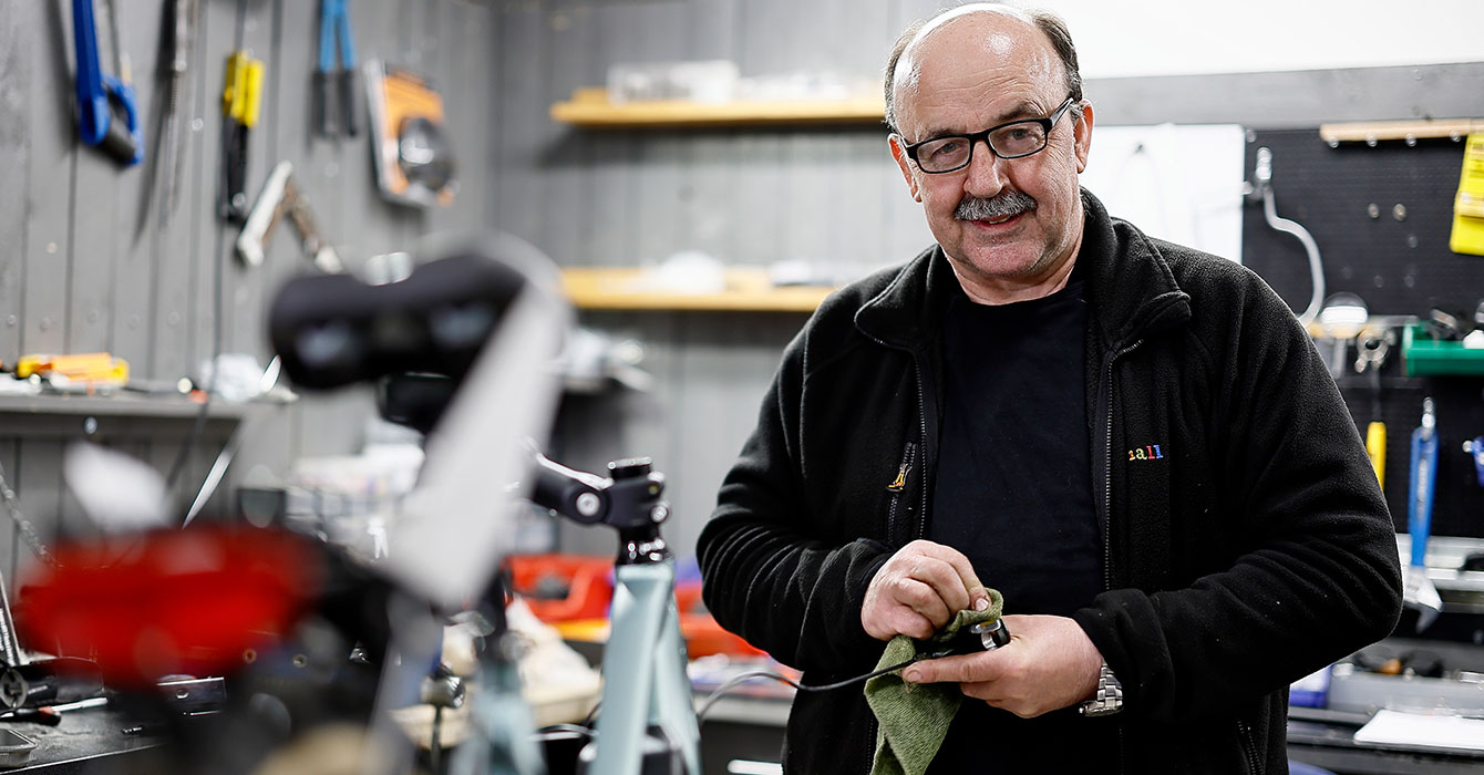 En man med glasögon står i en verkstad och rengör en liten komponent med en trasa. I bakgrunden syns verktyg och arbetsbänkar.