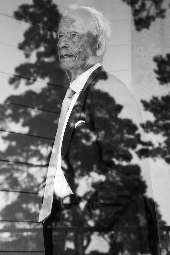 Svartvit bild av Björn i kostym och slips som står i profil med händerna i fickorna. Trädens skuggor faller över honom och skapar ett mönster av ljus och skugga över ansiktet och kavajen.