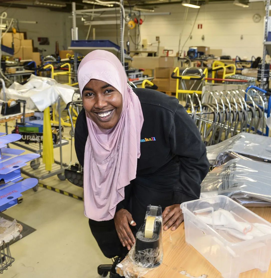 En leende kvinna med ljusrosa hijab står i en verkstadsmiljö med vagnar och utrustning i bakgrunden. Hon lutar sig över ett bord med en tejphållare och en plastlåda framför sig.