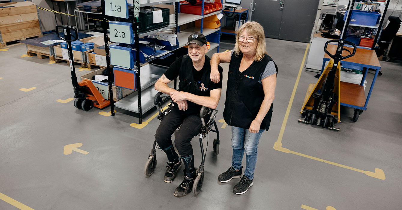 Bild på två medarbetare som står i ett lager. På golvet syns gula tejpmarkeringar. Medarbetarna är klädda i svart och tittar in i kameran. En står och en sitter i en rullstol. Bakom personerna står det hyllor med prylar.