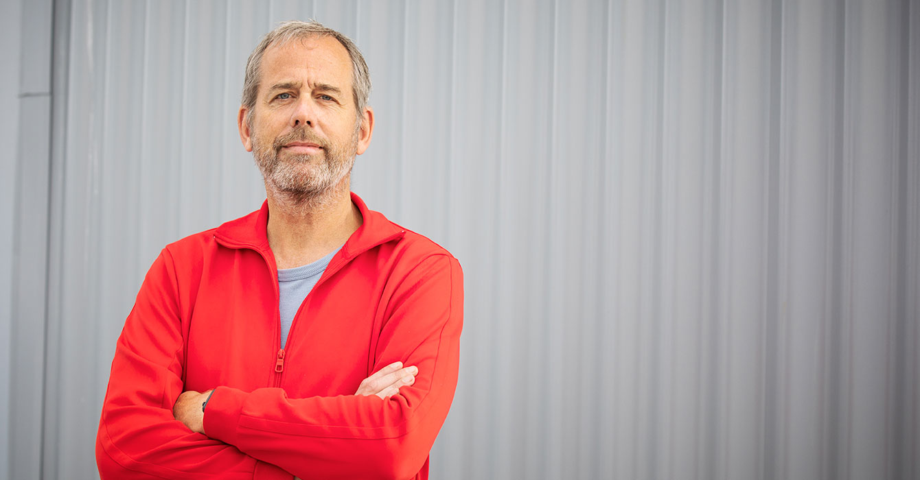 Jörgen Lorentzon med ljusbrunt hår och gråspräckligt skägg samt blå ögon står med armarna i kors och tittar självsäkert in i kameran framför en grå plåtvägg
