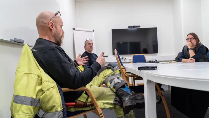 Kläddesignern Thomas Håkansson sitter vid ett bord och pratar med två män som bär arbetskläder. 