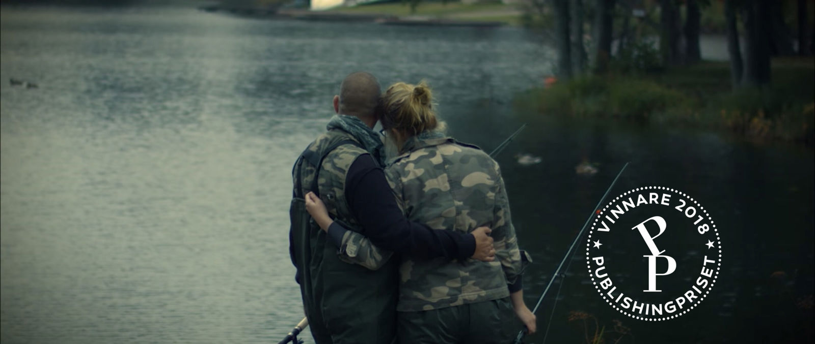 Två personer står med armarna om varandra vid en sjö och fiskar, med en symbol för vinnare av Publishingpriset 2018 i bilden.