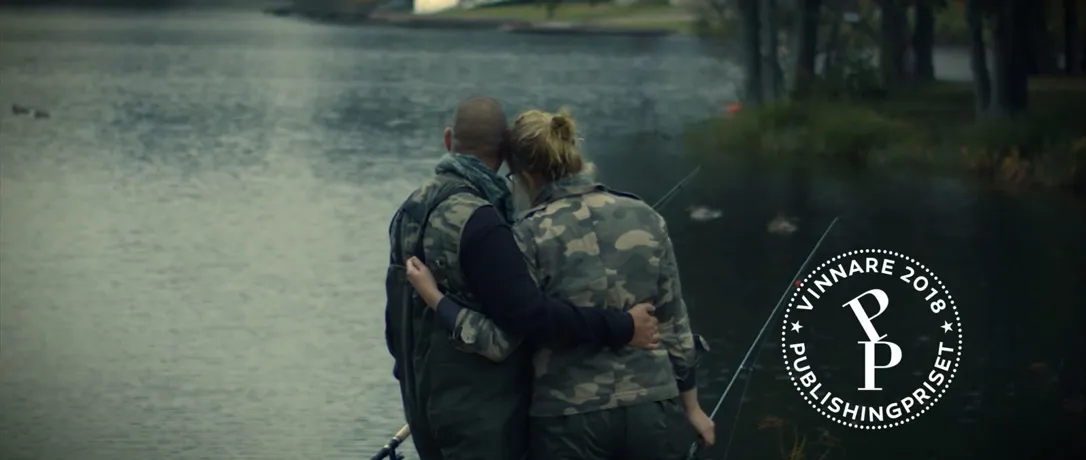 Två personer står med armarna om varandra vid en sjö och fiskar, med en symbol för vinnare av Publishingpriset 2018 i bilden.