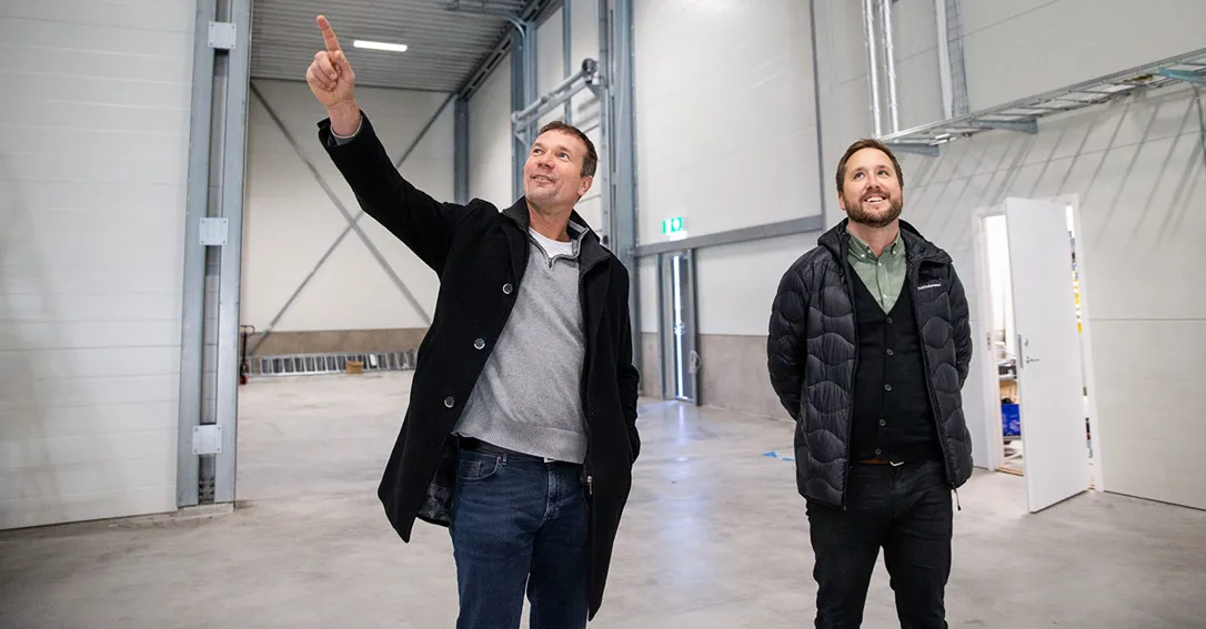 Två män i tom industrilokal. Mannen till vänster pekar med fingret åt taket. Båda tittar åt hållet mannen pekar.