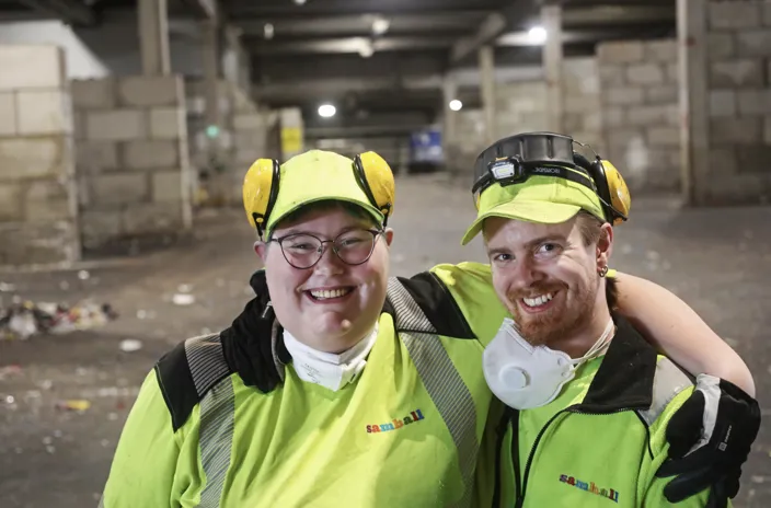 Jennifer Sjöberg och Eric Hjertz står bredvid varandra. De bär arbetskläder i reflexgult. De ler och ser in i kameran.