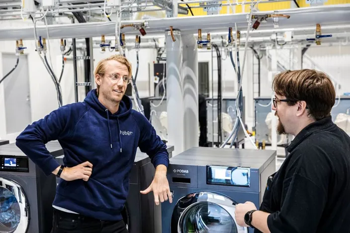 Två personer står och samtalar i en industrimiljö bredvid tvättmaskiner och rörinstallationer i taket. En av dem bär en mörkblå tröja med texten ”hyber”.