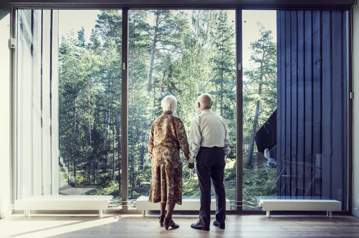 Lillemor och Björn står hand i hand framför ett stort panoramafönster och ser ut över en grön skog. De är fotograferade bakifrån, och dagsljuset strömmar in i rummet med trägolv och stora glaspartier.