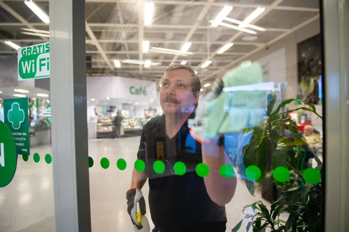 Alt-text: En man rengör en glasruta med en trasa i en butiksmiljö. På glaset sitter en skylt med texten ”Gratis WiFi”, och i bakgrunden syns ett caféområde.