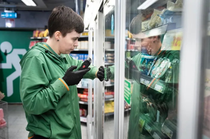 Nora Mångsén scannar varor i en kyl i butiken med ett verktyg. Hon bär gröna arbetskläder. 