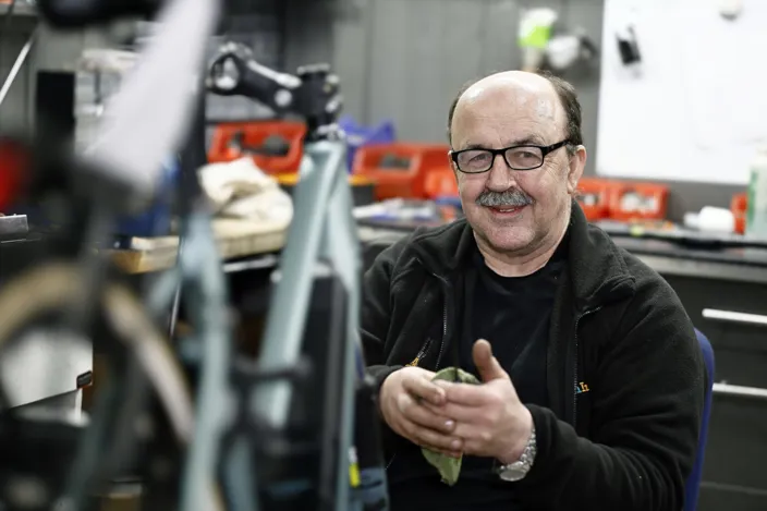 Mirsat Murjin sitter och mekar med en cykel i en verkstad. Han tittar in i kameran samtidigt som han torkar händerna med en trasa. 