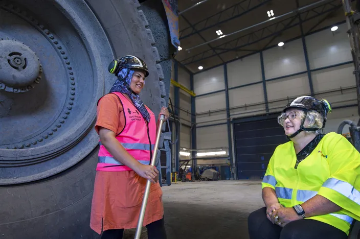 Gudrun sitter i en rullstol och bär en reflextröja och är ute på verkstadsgolvet med en medarbetare. Medarbetaren bär en rosa reflexväst. 