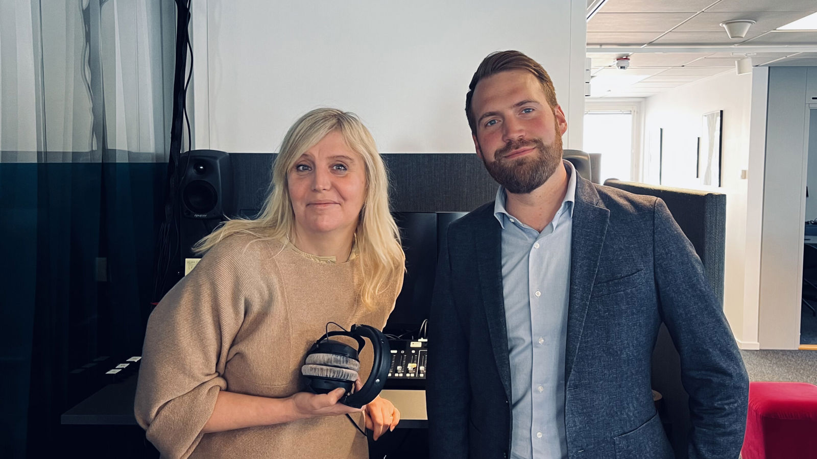 Två personer står i en studio- eller kontorsmiljö, där den ena håller i ett par hörlurar.