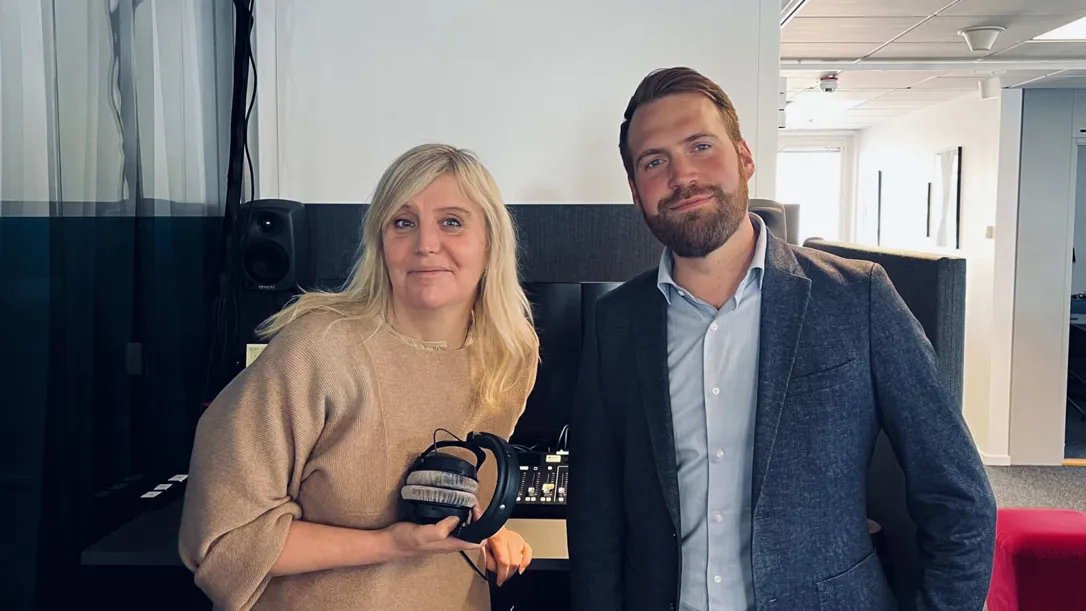 Två personer står i en studio- eller kontorsmiljö, där den ena håller i ett par hörlurar.