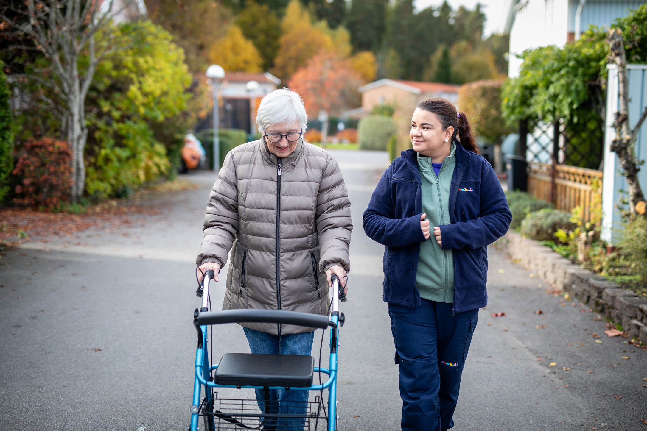 En Samhallmedarbetare promenerar tillsammans med en äldre kvinna som använder rollator i ett bostadsområde.