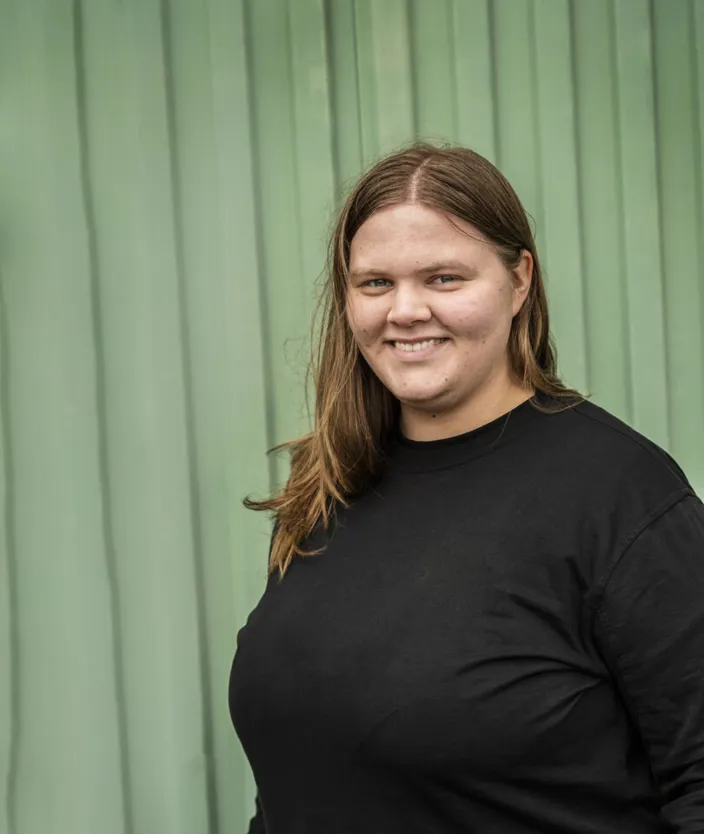 En ung kvinna med långt brunt hår ler mot kameran. Hon bär en svart tröja och står framför en grön vägg med vertikala paneler.