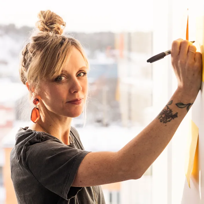 Kvinna tittar in i kameran och skriver på en whiteboard-tavla. 