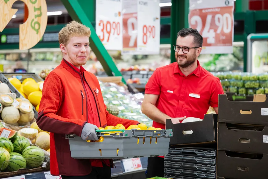 Två butiksanställda i röda tröjor arbetar i fruktavdelningen i en matbutik. En ung man bär en låda med citrusfrukter medan en äldre man i bakgrunden håller en melon. Prislappar och grönsaker syns i bakgrunden.