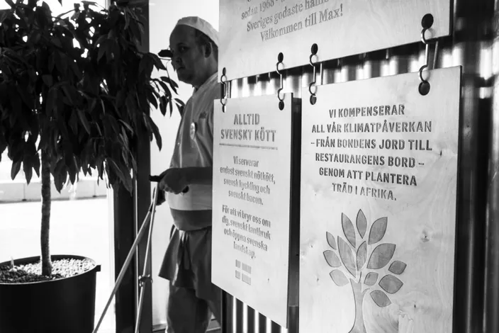 En svartvit bild av en restauranganställd i arbetskläder som står bredvid skyltar med information om svenskt kött och klimatkompensation genom trädplantering.
