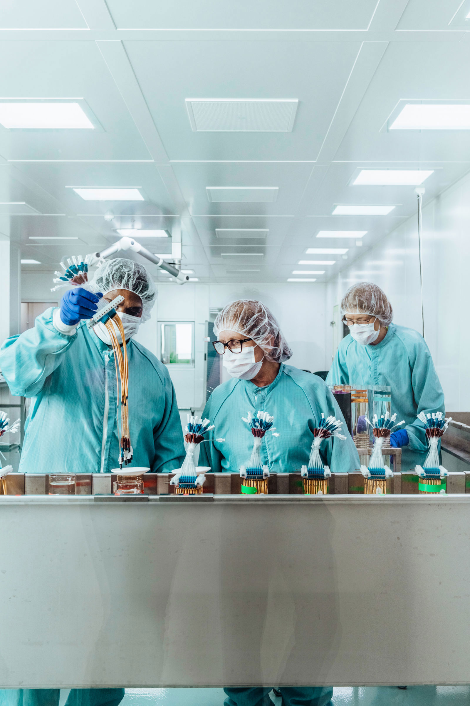 Tre personer i skyddskläder, munskydd och hårskydd arbetar i ett sterilt laboratorie- eller produktionsrum och hanterar små komponenter på ett löpande band.
