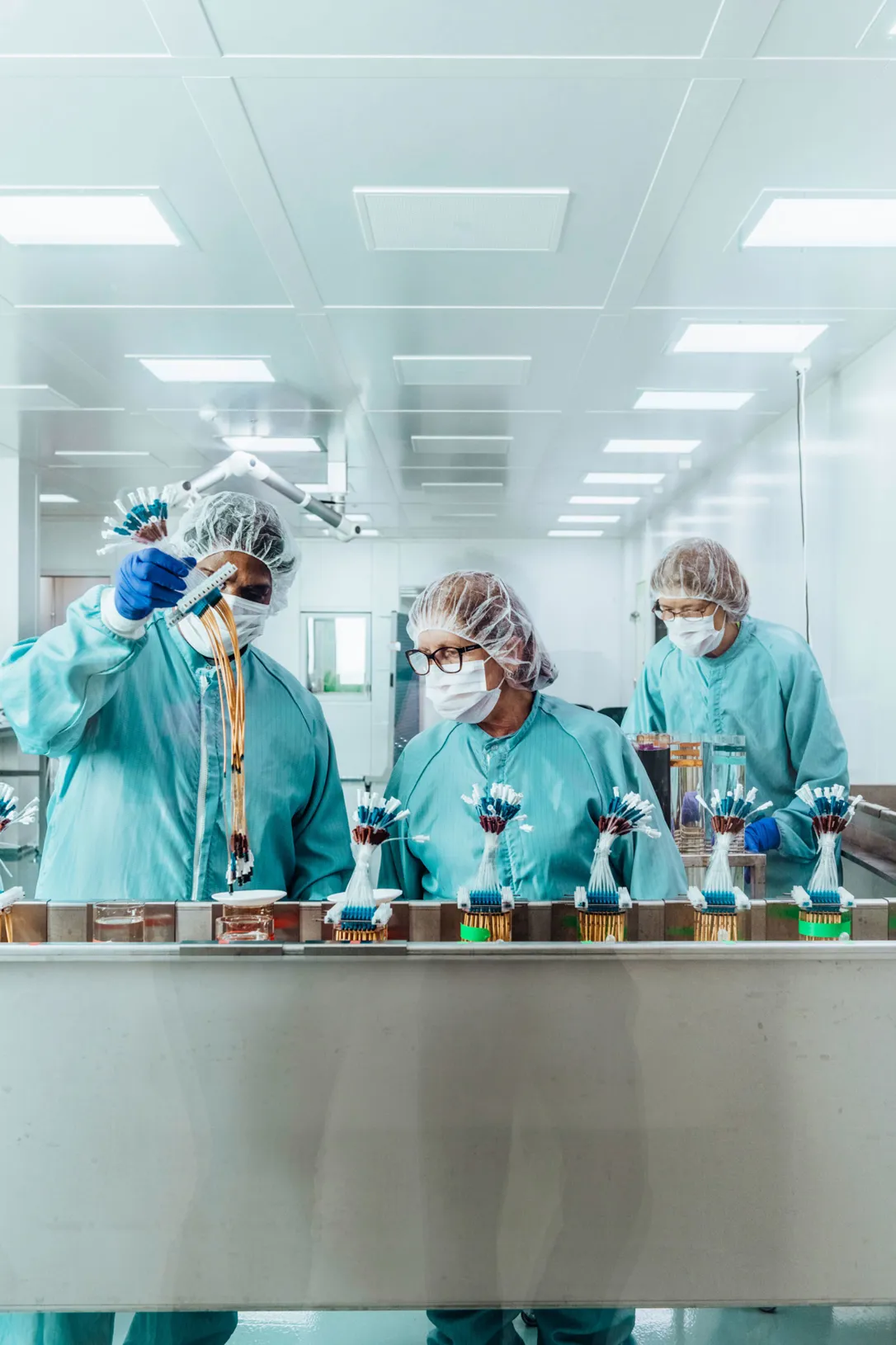 Tre personer i skyddskläder, munskydd och hårskydd arbetar i ett sterilt laboratorie- eller produktionsrum och hanterar små komponenter på ett löpande band.