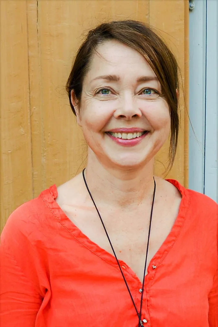 Tatja Hirvikoski ler och tittar in i kameran. Hon bär en röd topp och står framför en gul vägg.
