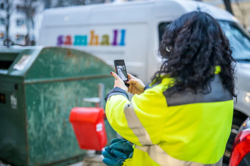 En person i reflexjacka fotograferar ett grönt sopkärl med sin mobiltelefon, bakom syns en vit skåpbil med texten "samhall".