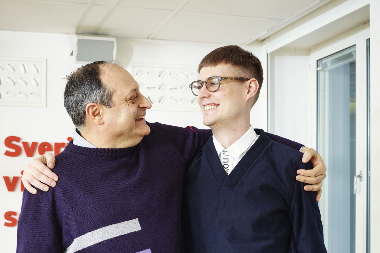 Två personer står nära varandra och ler medan de håller om varandras axlar i en inomhusmiljö.