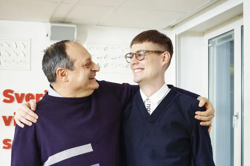 Två personer står nära varandra och ler medan de håller om varandras axlar i en inomhusmiljö.