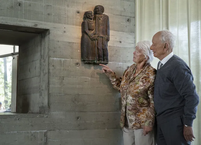 Lillemor och Björn står vid en betongvägg inomhus och tittar på en väggmonterad träskulptur som föreställer ett par.
