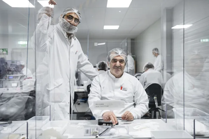 Bild på två personer i vita rockar och nätskydd på håret. De är omringdade av kollegor med samma arbetskläder. Rummet är vitt och arbetsytorna är avgränsade med glasrutor.