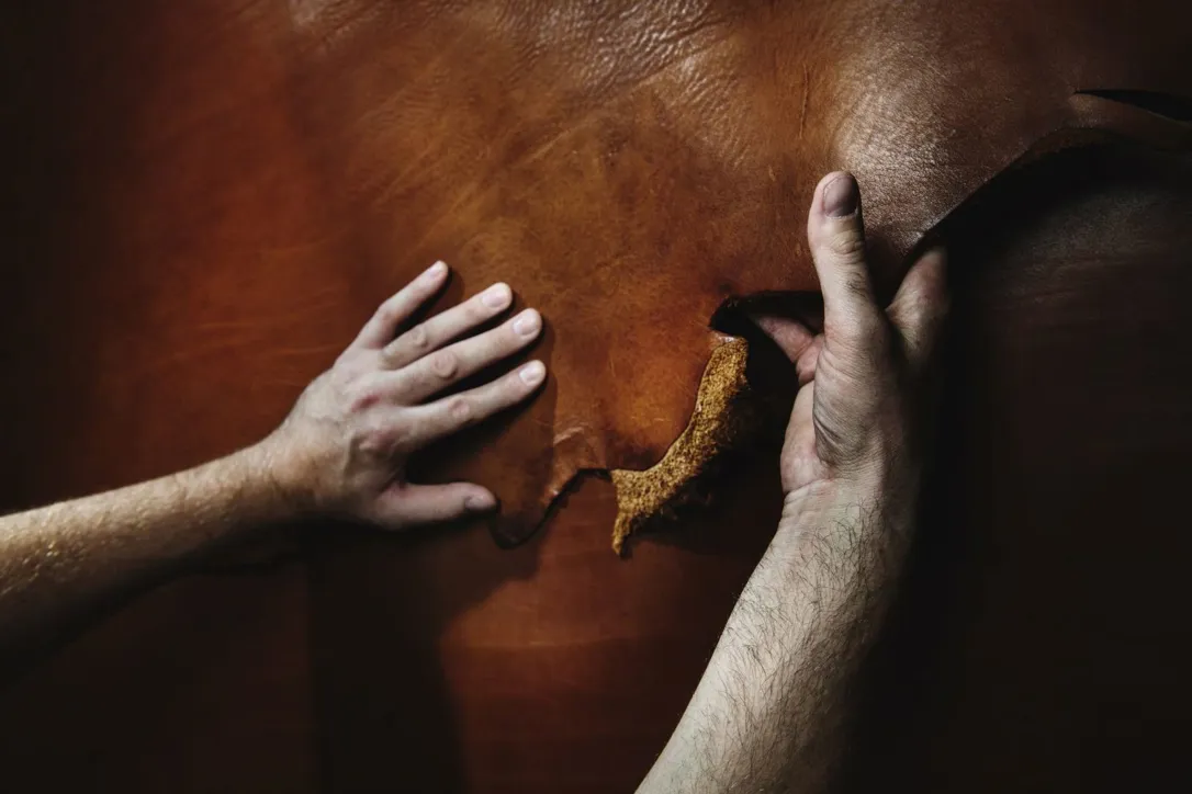 Två händer håller i och lyfter en flik av brunt läder, så att den mjuka undersidan syns. Närbilden visar materialets struktur och yta i detalj.