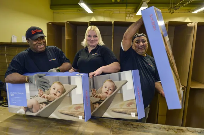 Tre lagerarbetare står framför staplade kartonger och håller stora produktförpackningar med en bild av en bebis i en babysitter. De ler och poserar med kartongerna i en lagerlokal.