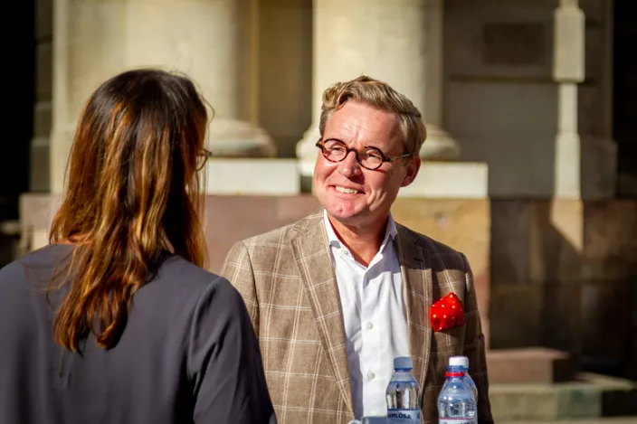 En man i kavaj med röd näsduk och glasögon står utomhus och samtalar med en kvinna. I förgrunden syns två vattenflaskor och i bakgrunden skymtar en byggnad i solljus.