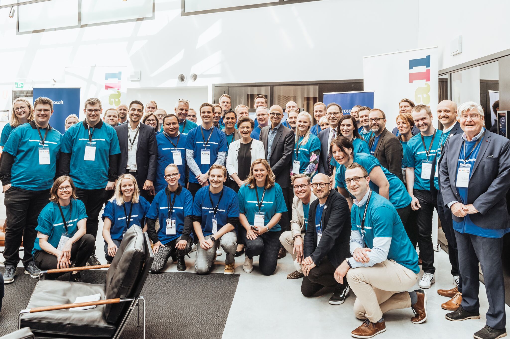 En stor grupp deltagare i blå t-shirts och namnskyltar poserar tillsammans för ett gruppfoto vid ett evenemang inomhus, med rollups och konferensmiljö i bakgrunden.