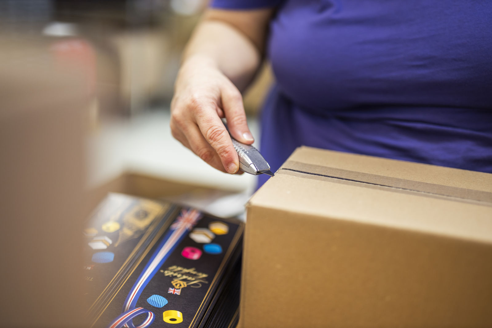 Närbild på en samhallsmedarbetare som öppnar en kartong med en kartongkniv vid ett packbord.