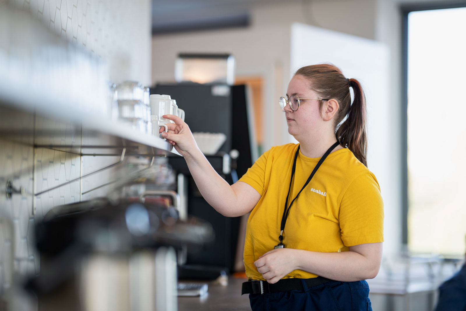 Samhallsmedarbetare i gul tröja ställer upp koppar på en hylla i köksmiljö.