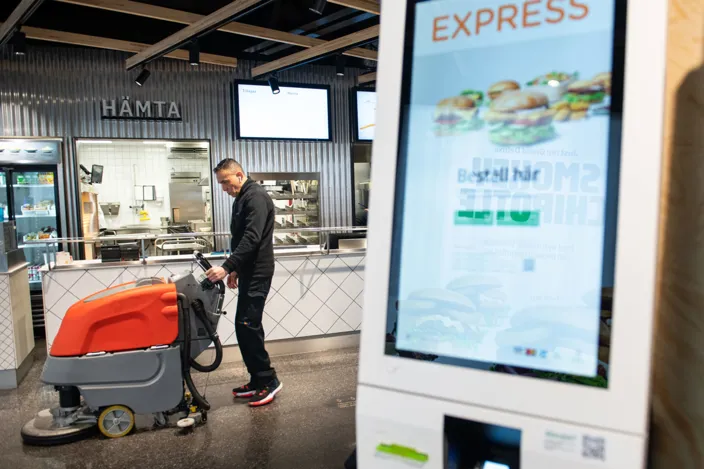 Humberto Rengifo går med en sopmaskin och tvättar golvet i en max-restaurang. I förgrunden syns en beställnignsmaskin. 