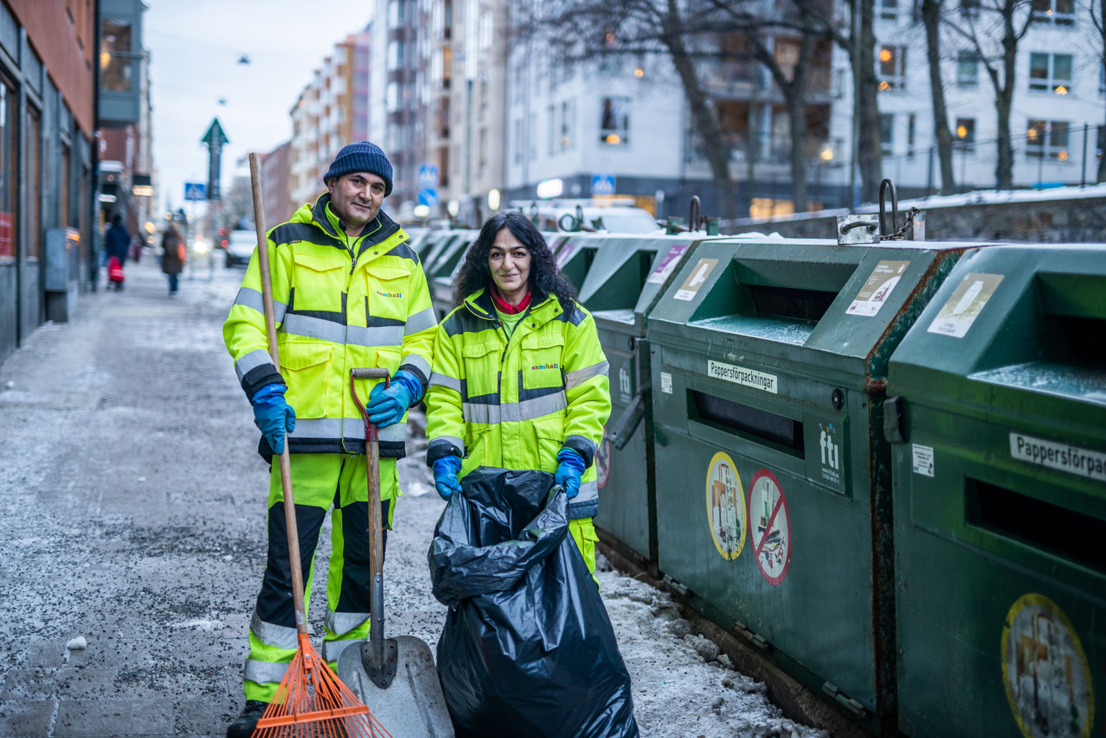 Två personer i varselkläder står vid återvinningskärl och håller i städredskap och sopsäck.