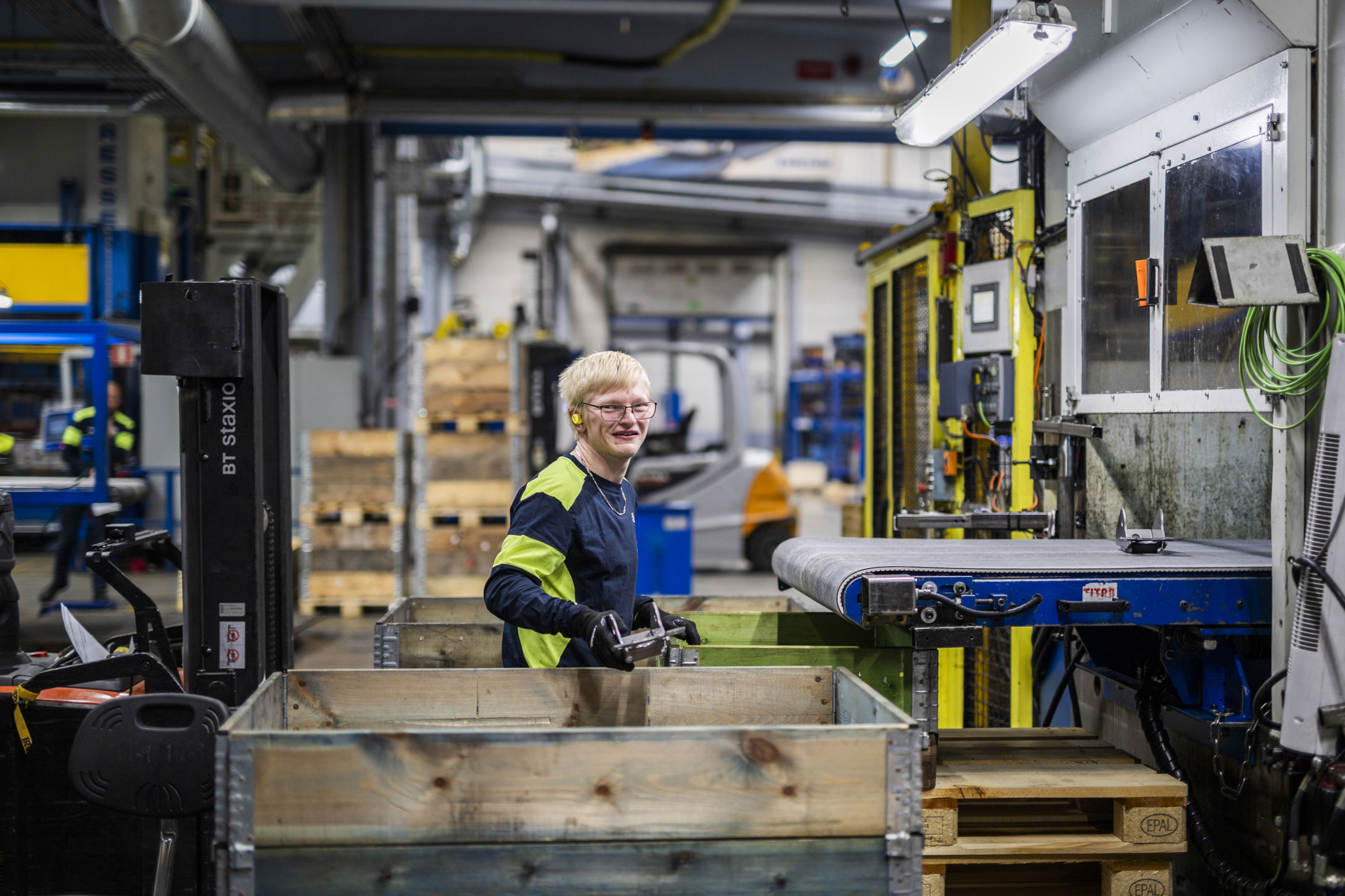 En Samhallmedarbetare arbetar vid en maskin i en industrimiljö och hanterar material i en trälåda.
