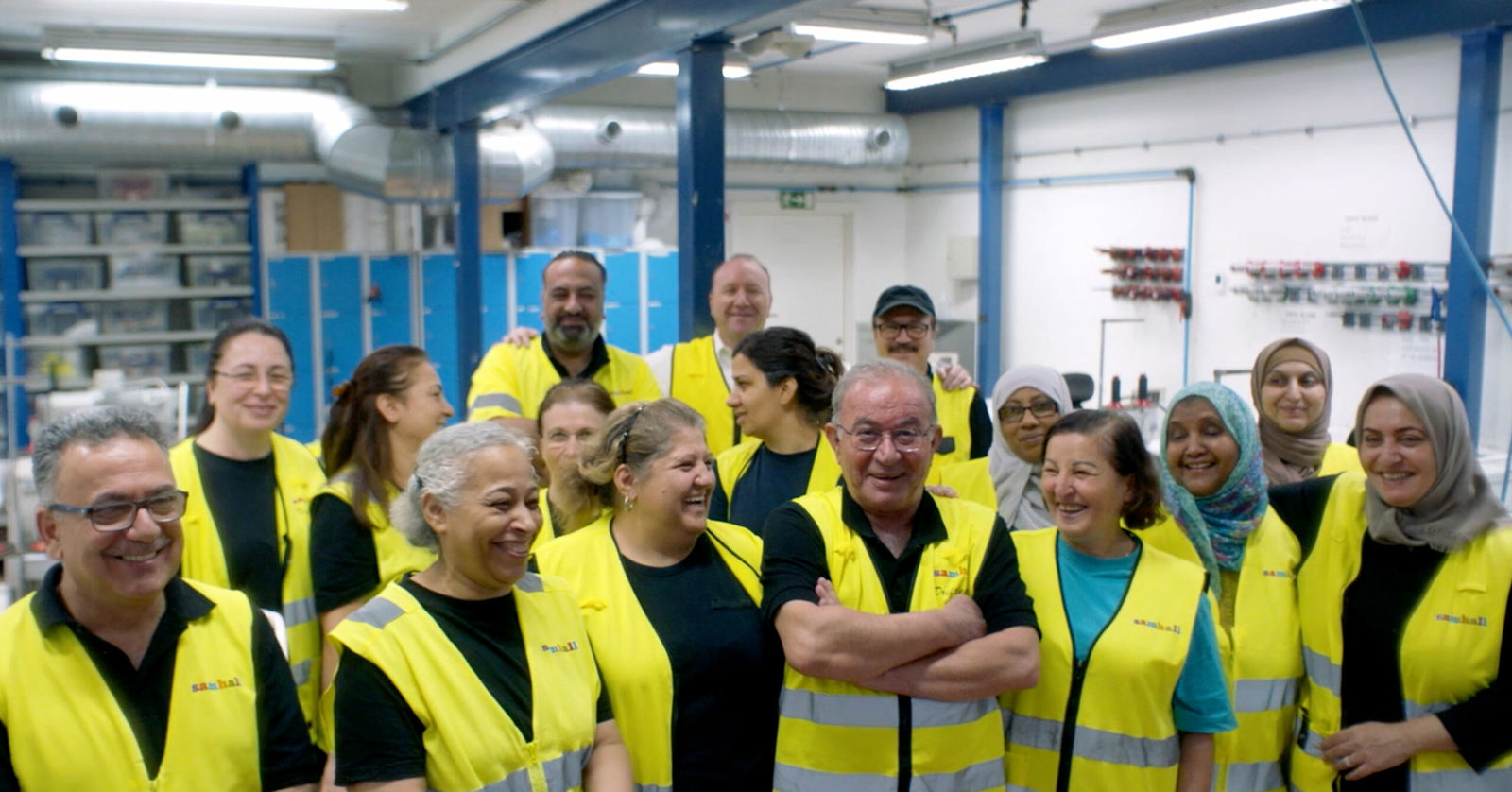 En grupp medarbetare i gula varselvästar står tillsammans i en industrilokal och skrattar, med ventilationsrör och arbetsutrustning i bakgrunden.