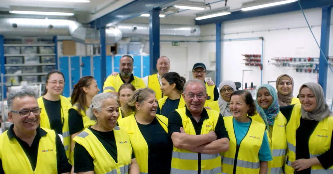 En grupp medarbetare i gula varselvästar står tillsammans i en industrilokal och skrattar, med ventilationsrör och arbetsutrustning i bakgrunden.