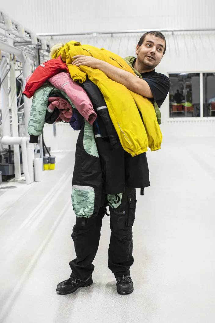 En man står i en ljus industrilokal och bär en stor bunt färgglada jackor över axeln, bland annat en gul, röd och rosa jacka.