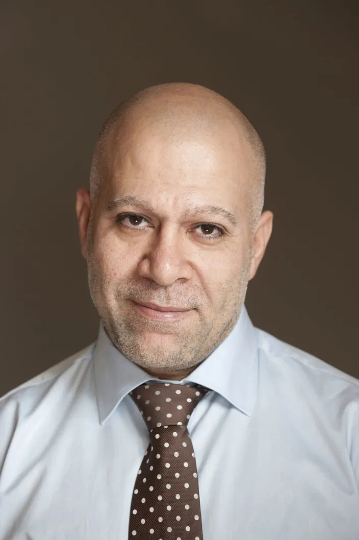 Porträtt av man som tittar in i kameran. Han har inget hår på huvudet och bär en brun slips med vita prickar och ljus skjorta. 