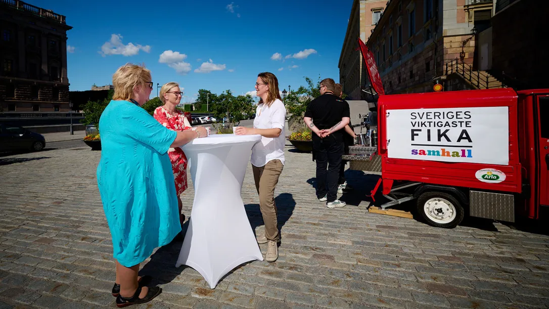 Personer står runt ett bord ute i solen bredvid en fikavagn