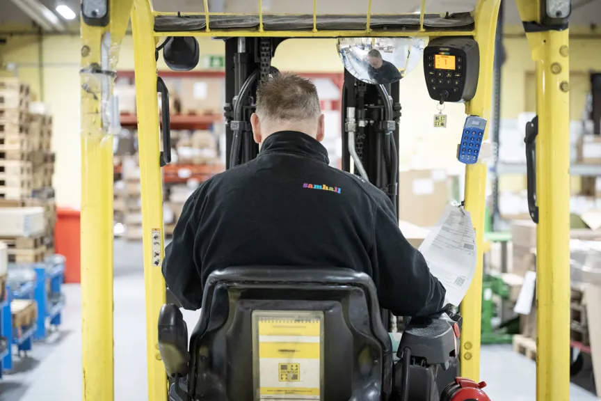 Samhall-medarbetare kör en gaffeltruck i ett lager fullt med kartonger och pallar.